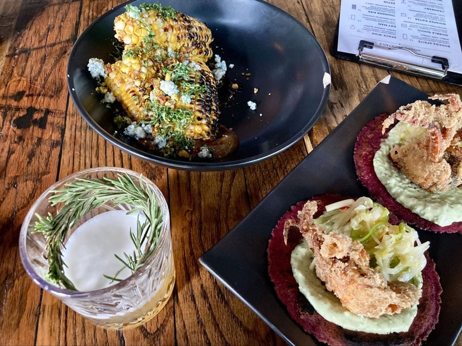 Una copa de cóctel de ginebra Whisper Sister, costillas de maíz y tacos de cangrejo de caparazón blando en una mesa en Misfit Snack Bar